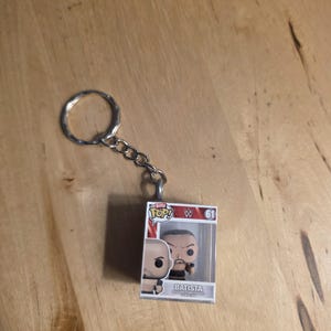 May include: A Funko Pop! keychain featuring a Batista figure in a clear plastic case. The keychain includes a silver metal ring and chain. The figure is in a box with the words "POP!" and "BATISTA" printed on it.