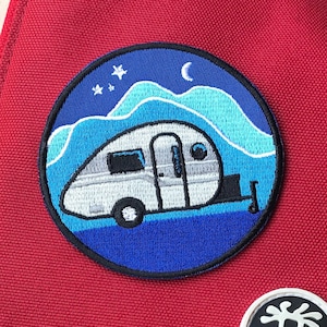 May include: A red messenger bag with a blue and white embroidered patch of a camper van in front of a mountain scene. The patch also includes a crescent moon and stars. The bag also has a white embroidered patch of a stick figure with a circle around it.