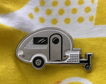 May include: A silver enamel pin of a small, white camper trailer with black details. The camper has a window, a door, and a tire. The pin is on a yellow and white patterned fabric.