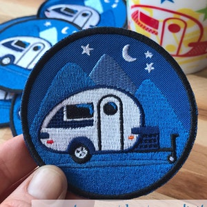 Könnte beinhalten: Ein blauer gestickter Aufnäher mit einem weißen Wohnwagen, Bergen, einer Mondsichel und Sternen. Der Aufnäher ist rund und hat einen schwarzen Rand. Der Text "www.etsy.com/shop/sarandpitous" ist unten im Bild sichtbar.