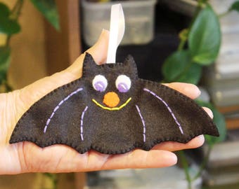 May include: A black felt bat with purple stitching and a smiling face. The bat has white eyes with purple pupils and an orange nose. The bat is hanging from a white ribbon.