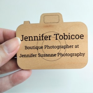 May include: A wooden business card shaped like a camera. The card has the name "Jennifer Tobicoe" and "Boutique Photographer at Jennifer Susanne Photography" printed on it. The card is light brown with black text.