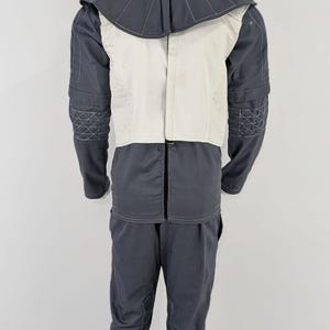 May include: A two-piece grey jumpsuit with a white vest and a high collar. The collar and sleeves have decorative stitching. The trousers have a straight leg design. The outfit is displayed on a mannequin.