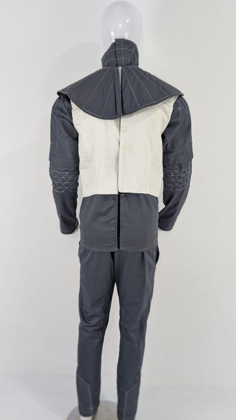 May include: A two-piece grey jumpsuit with a white vest and a high collar. The collar and sleeves have decorative stitching. The trousers have a straight leg design. The outfit is displayed on a mannequin.