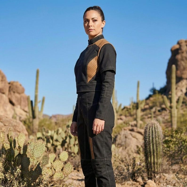 May include: A person wearing a black jumpsuit with brown accents on the shoulders and legs, standing in a desert landscape with cacti and mountains under a clear blue sky. The jumpsuit has a high collar and long sleeves.