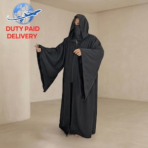 Peut inclure: Une longue robe noire à capuche avec de larges manches et un voile facial. La robe est ouverte sur le devant et descend jusqu'au sol. Les mots "DUTY PAID DELIVERY" sont imprimés en rouge en haut à gauche de l'image.