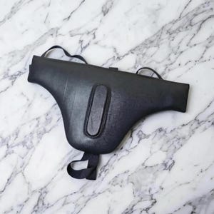 May include: A black leather saddle bag with a unique, curved shape. The bag features a long, oval-shaped accent on the front and a strap for carrying. The bag is shown on a white marble surface.