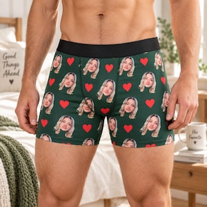 Op de afbeelding: Donkergroene boxershort met een herhalend patroon van een gezicht en rode hartjes. Het kledingstuk heeft een effen zwarte elastische tailleband. Het ontwerp bestaat uit meerdere exemplaren van hetzelfde gezicht met lang blond haar.