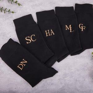 Puede incluir: Cinco calcetines negros con iniciales doradas bordadas se muestran sobre una superficie gris. Las iniciales son DN, SC, HA, ML y GF. Los calcetines están doblados.