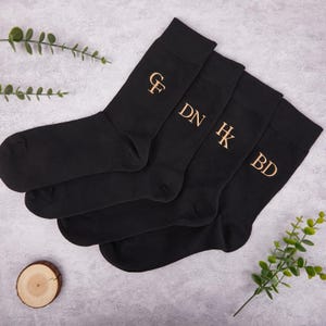 May include: Four pairs of black socks with gold embroidered initials: GF, DN, HK, and BD. The socks are arranged on a light gray surface with decorative greenery and a small wooden slice. The socks are a dark, solid color.