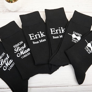 May include: Set of black socks with white text. The socks have different names and the words "Best Man" and "Groom" printed on them. The socks also have a date printed on them.