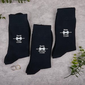 Può includere: Tre paia di calzini blu navy con testo bianco e grafica a farfallino. Ogni calzino è personalizzato con un nome e un ruolo: David, Sposo; Kevin, L'Ufficiale; e Brian, Testimone, insieme a una data. Tema matrimonio.