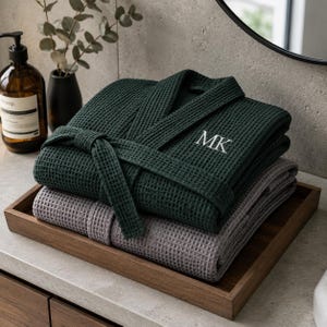 May include: A dark green waffle-weave robe with the embroidered letters MK on the chest, folded neatly on top of a light grey waffle-weave robe. Both robes feature a matching tie belt and a textured grid pattern, stacked together on a wooden tray.