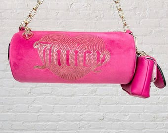 May include: A bright pink, cylindrical handbag with a gold chain strap and a smaller matching pouch. The bag features a rhinestone-studded heart design with the word "Juicy" in gold lettering.