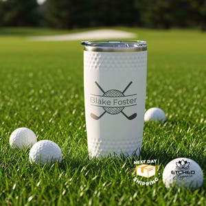 May include: White golf-themed tumbler with a silver lid, featuring a golf ball and crossed clubs design, and the name "Blake Foster." The tumbler is surrounded by golf balls on a green grassy field.