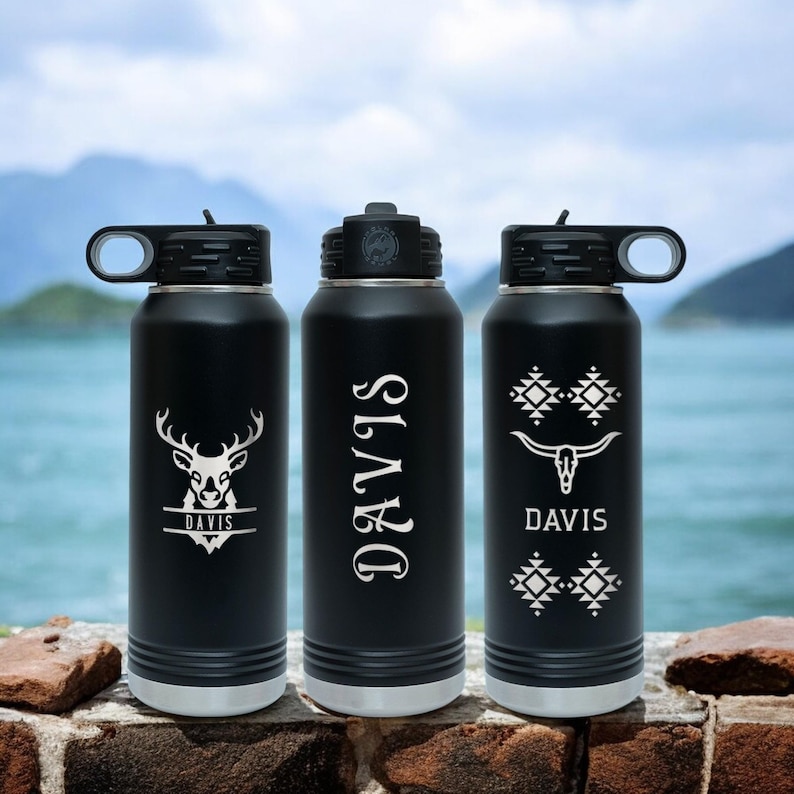May include: Three black stainless steel water bottles with different designs. The first bottle has a deer head with the name "DAVIS" underneath. The second bottle has the name "DAVIS" in a vertical script font. The third bottle has a skull with horns and a geometric pattern with the name "DAVIS" underneath.