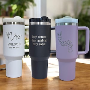May include: Three stainless steel travel mugs with different engravings. The white mug says "Mrs. WILSON est 2024". The black mug says "Very demure Very mindful Very sober". The purple mug says "Tinsle Bella" with two bunny illustrations.