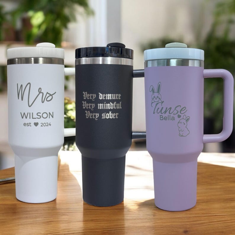 May include: Three stainless steel travel mugs with different engravings. The white mug says "Mrs. WILSON est 2024". The black mug says "Very demure Very mindful Very sober". The purple mug says "Tinsle Bella" with two bunny illustrations.