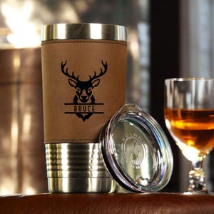 May include: Personalized stainless steel travel mug with a brown leather wrap. The mug features a deer head design and the name "Bruce" engraved on a split line. The mug comes with a clear lid.