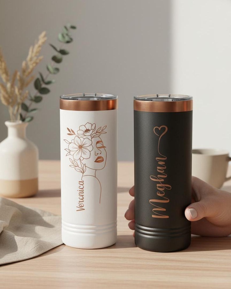 May include: Two insulated tumblers. One is white with a floral design and the name "Veronica" in a copper colour. The other is black with the name "Meghan" and a heart in copper. Both have copper-coloured rims.
