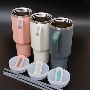 May include: Three stainless steel travel mugs with lids and straws. The mugs are in pink, white, and gray. Each mug has a handle with a matching color accent.