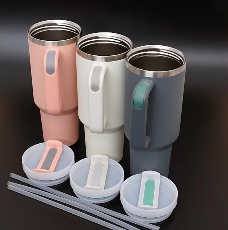 May include: Three stainless steel travel mugs with lids and straws. The mugs are in pink, white, and gray. Each mug has a handle with a matching color accent.