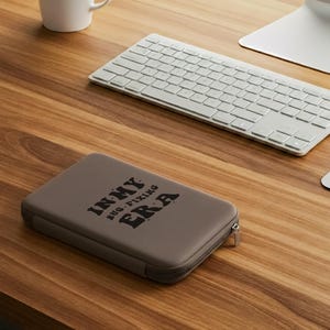 May include: A taupe-colored zippered case with the text "IN MY BUG-FIXING ERA" in black lettering. The case is rectangular and sits on a wooden desk next to a white keyboard and a computer monitor.