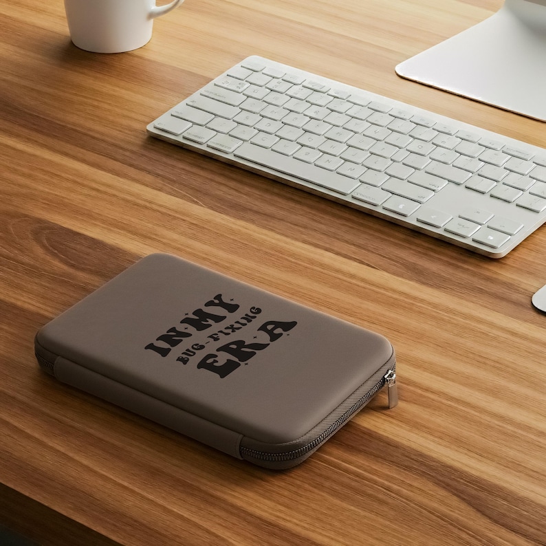 May include: A taupe-colored zippered case with the text "IN MY BUG-FIXING ERA" in black lettering. The case is rectangular and sits on a wooden desk next to a white keyboard and a computer monitor.