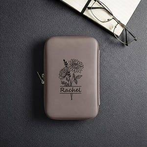 May include: A gray, rectangular case with a zipper closure, featuring a black floral design and the name "Rachel" underneath. The case is on a dark gray surface, with a notebook and glasses in the background.