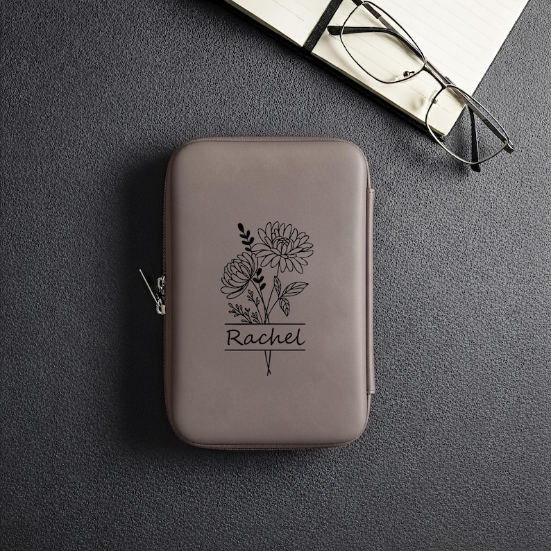 May include: A gray, rectangular case with a zipper closure, featuring a black floral design and the name "Rachel" underneath. The case is on a dark gray surface, with a notebook and glasses in the background.