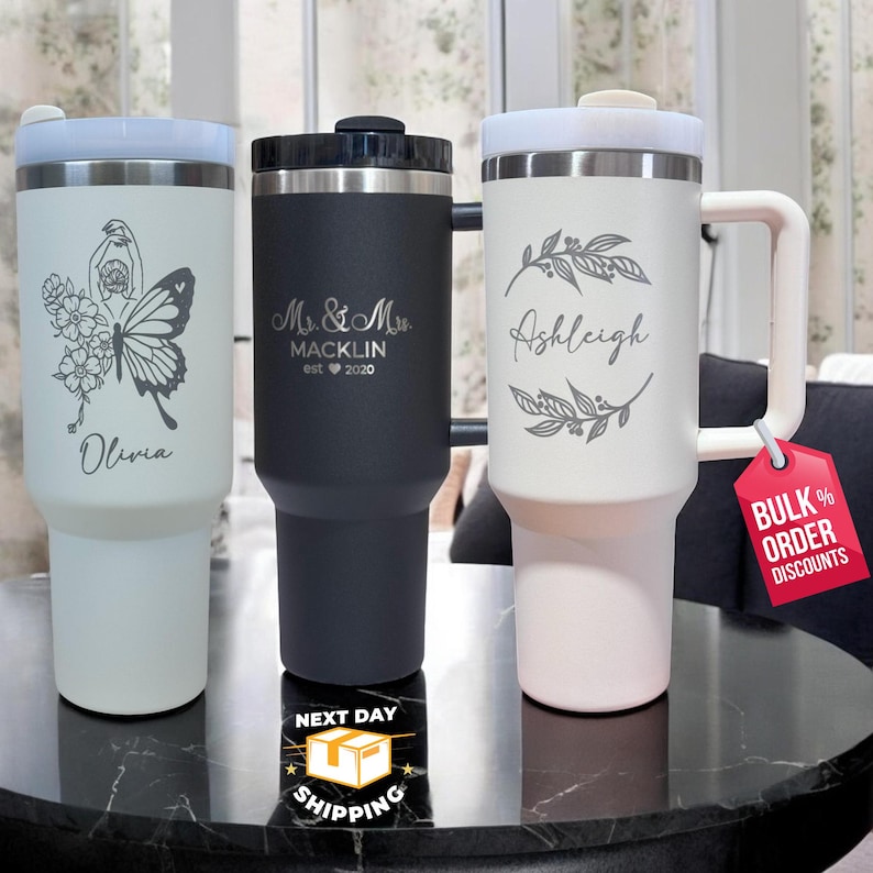May include: Three stainless steel travel mugs with different designs. The first mug is white with a butterfly and floral design and the text "Olivia". The second mug is black with the text "Mr. & Mrs. Macklin est 2020". The third mug is white with a floral design and the text "Ashleigh".