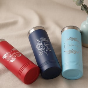 May include: Three personalised tumblers in red, navy blue, and light blue. Each tumbler has a silver lid and engraved text: "Octavia" with a butterfly, "Jennifer" with a floral design, and "Victoria" with leaves. The tumblers are cylindrical.