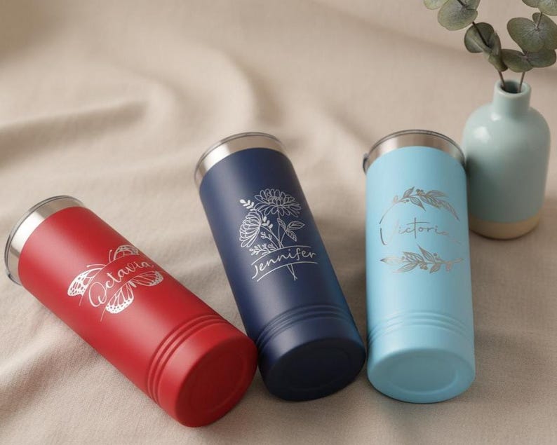 May include: Three personalised tumblers in red, navy blue, and light blue. Each tumbler has a silver lid and engraved text: "Octavia" with a butterfly, "Jennifer" with a floral design, and "Victoria" with leaves. The tumblers are cylindrical.