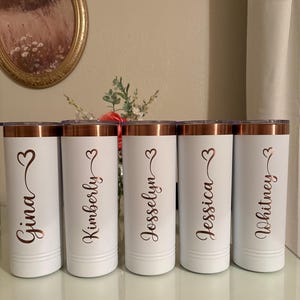 May include: Five white insulated tumblers with copper-coloured rims and lids. Each tumbler is personalised with a name in a copper-coloured script font and a heart symbol. The names are Gina, Kimberly, Jesselyn, Jessica, and Whitney.