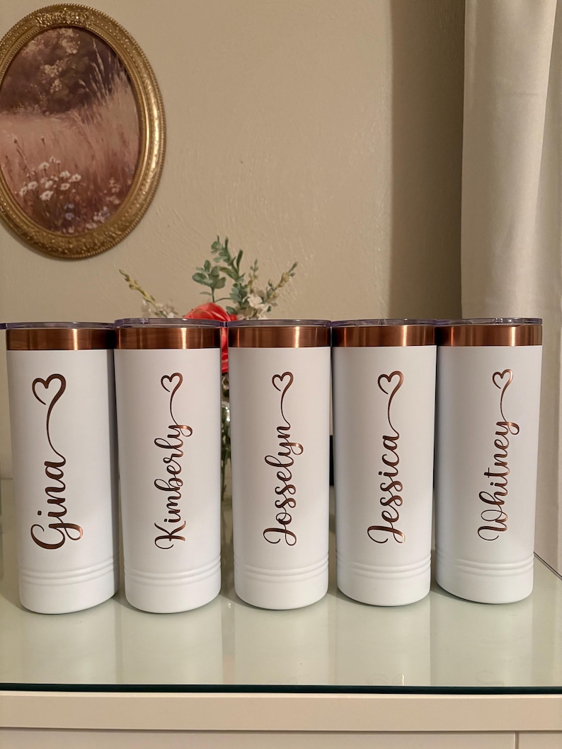 May include: Five white insulated tumblers with copper-coloured rims and lids. Each tumbler is personalised with a name in a copper-coloured script font and a heart symbol. The names are Gina, Kimberly, Jesselyn, Jessica, and Whitney.