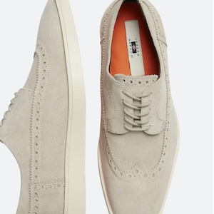 Könnte beinhalten: Ein Paar hellgraue Wildlederschuhe im Brogue-Stil mit weißen Sohlen. Die Schuhe haben einen Schnürverschluss und ein perforiertes Design. Der Markenname "JOSEPH ABBOUD" ist auf der Innenseite des rechten Schuhs sichtbar.