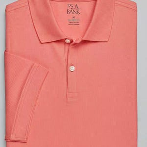May include: A coral pink polo shirt with a J.S.A. Bank label. The shirt is made of 100% cotton and is made in Cambodia.