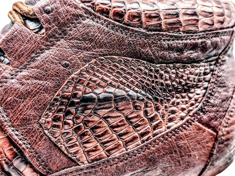 May include: Close-up of a brown leather shoe with a textured, crocodile-like pattern. The shoe features intricate stitching and a lace-up design. The leather has a rich, reddish-brown hue, with darker accents.