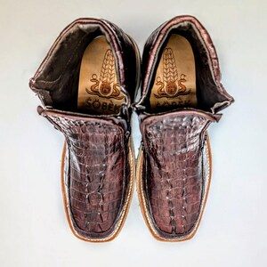 May include: A pair of brown leather boots with a crocodile pattern. The boots have a zipper on the side and a brown leather sole. The boots have a label on the inside that says "Sobek".