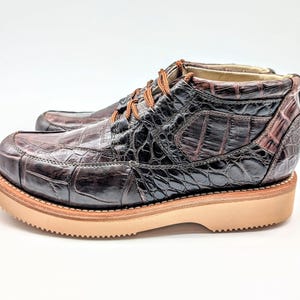 May include: A pair of brown leather boots with a crocodile skin pattern. The boots have a rounded toe, tan soles, and brown laces. The boots are approximately 6 inches tall.