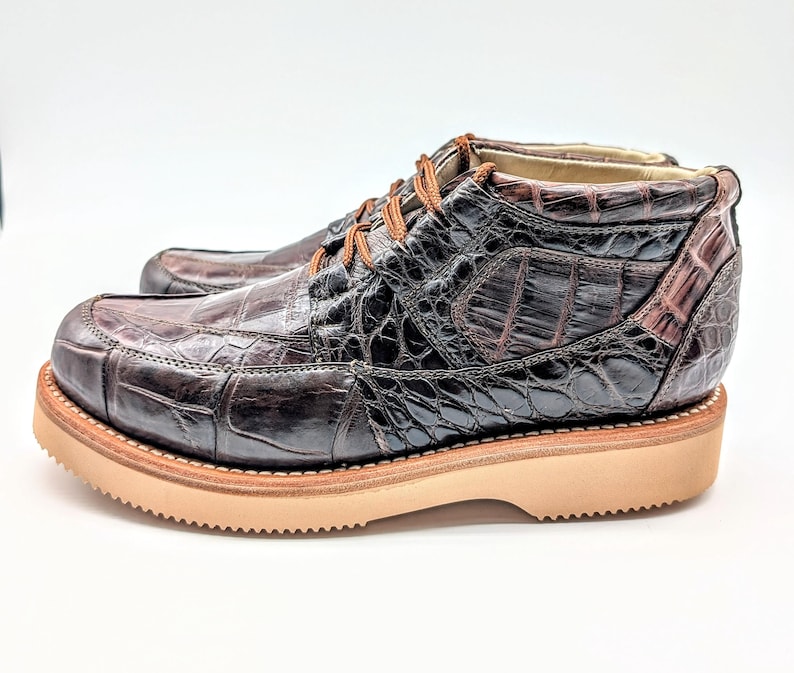 May include: A pair of brown leather boots with a crocodile skin pattern. The boots have a rounded toe, tan soles, and brown laces. The boots are approximately 6 inches tall.