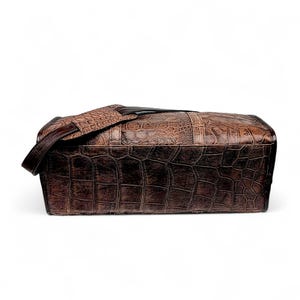 May include: A brown leather satchel bag with a crocodile skin texture. The bag has a shoulder strap and a rectangular shape. The color is a mix of dark and light browns, with a textured surface. The bag appears to be made of high-quality leather.