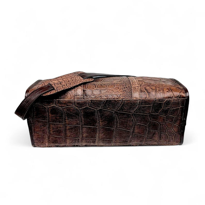May include: A brown leather satchel bag with a crocodile skin texture. The bag has a shoulder strap and a rectangular shape. The color is a mix of dark and light browns, with a textured surface. The bag appears to be made of high-quality leather.