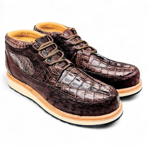 May include: A pair of brown leather ankle boots with a crocodile skin texture on the upper and toe. The boots have a light-coloured sole and brown laces. The interior lining is a light tan colour.