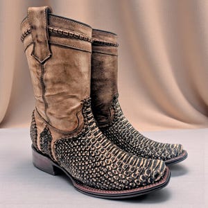 Könnte beinhalten: Ein Paar braune und schwarze Cowboy-Stiefel. Die Stiefel haben ein strukturiertes, schuppenartiges Muster am unteren Teil und ein glattes, hellbraunes Leder am oberen Teil. Sie haben eine quadratische Spitze und einen gestapelten Absatz.