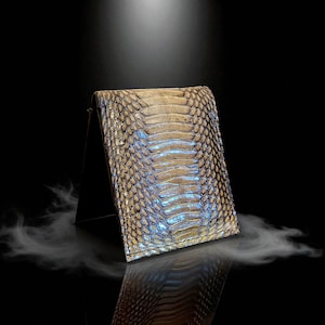 Op de afbeelding: Een bi-fold portemonnee met een slangenleerpatroon in tinten beige, bruin en blauw. De portemonnee wordt gepresenteerd op een zwarte standaard tegen een donkere achtergrond, met een reflecterend oppervlak eronder.