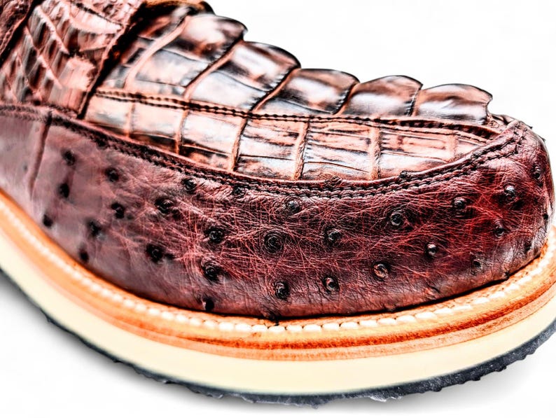 May include: Close-up of a brown leather shoe. The upper features a crocodile pattern, while the sides have an ostrich leather texture. The shoe has a cream sole with a black outsole and orange trim.