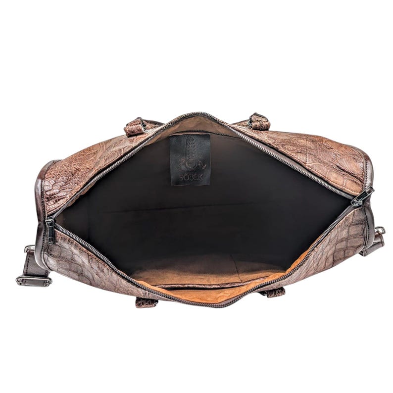 May include: Brown leather duffel bag with a textured crocodile pattern. The bag is open, revealing a black interior. The zipper is silver. A black tag with the text "SOULK" is visible inside the bag.