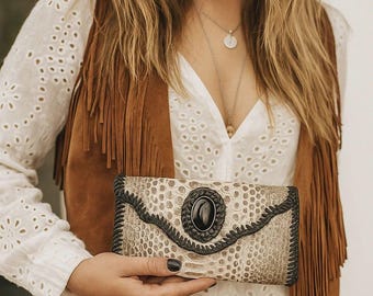 May include: A beige and black clutch bag with a snake-skin pattern and a large black oval stone. The clutch is held in front of a white eyelet shirt and a brown suede fringe vest. The overall aesthetic is bohemian.