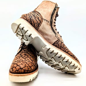 May include: Brown and tan leather boots with a unique textured pattern. The boots have a side zipper, lace-up closure, and a thick white sole. The soles have a tread pattern for grip. The boots are set against a white background.